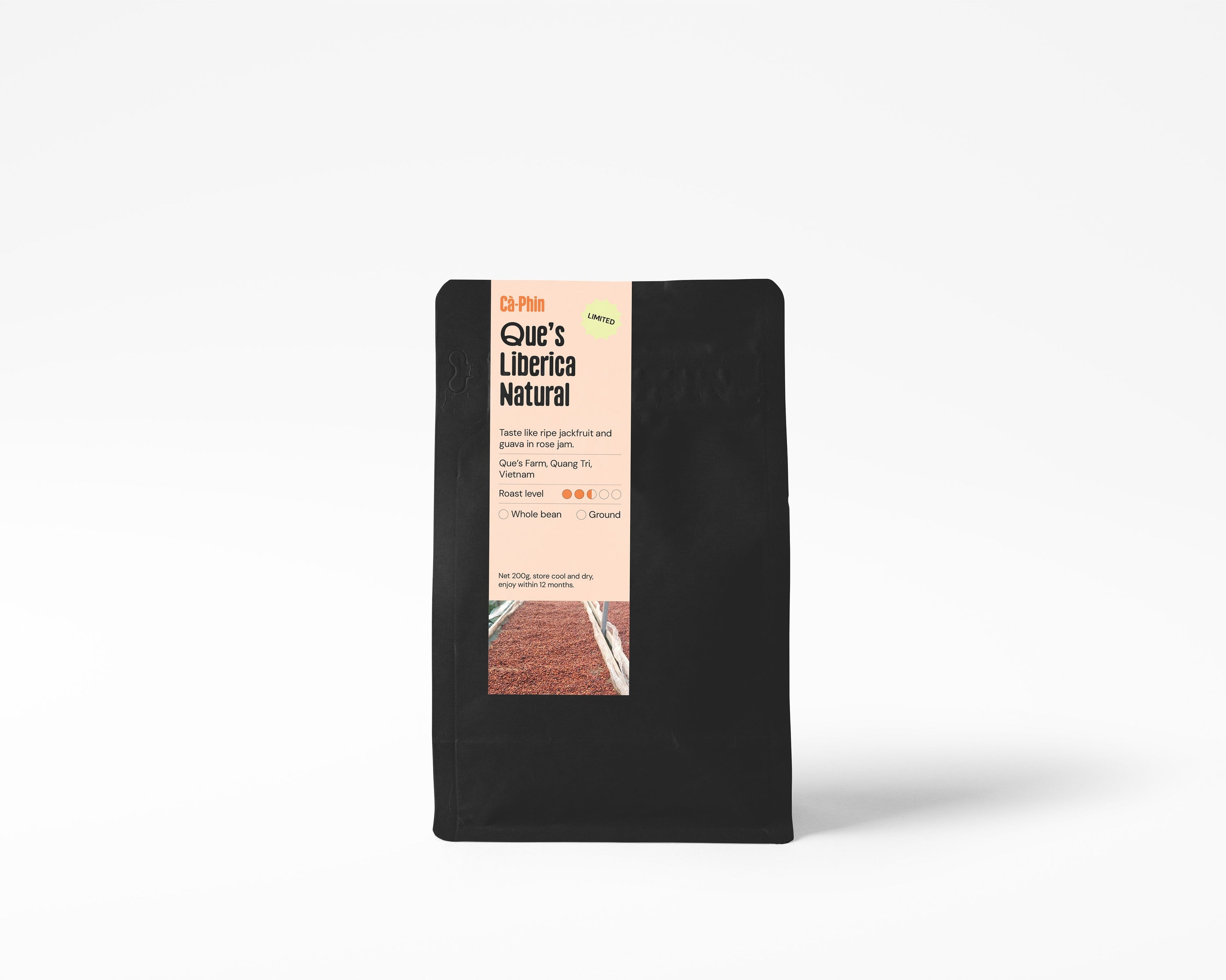 A black bag of Que's Liberica Natural coffee stands upright against a clean white background. The packaging features a vertical label with a peach background, black text, and a small image of coffee beans at the bottom.